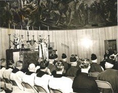Bishop Sheen at the altar during Mass in the parish hall, a large mural behind him and the congregation seated