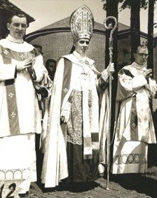 Bishop Sheen in mitre and cope, holding his crozier, between two vested deacons outside a brick building