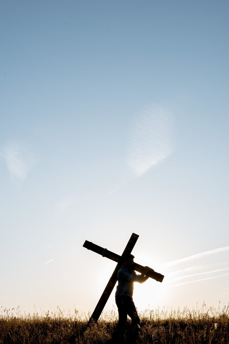 A man carrying a wooden cross through a field at sunrise
