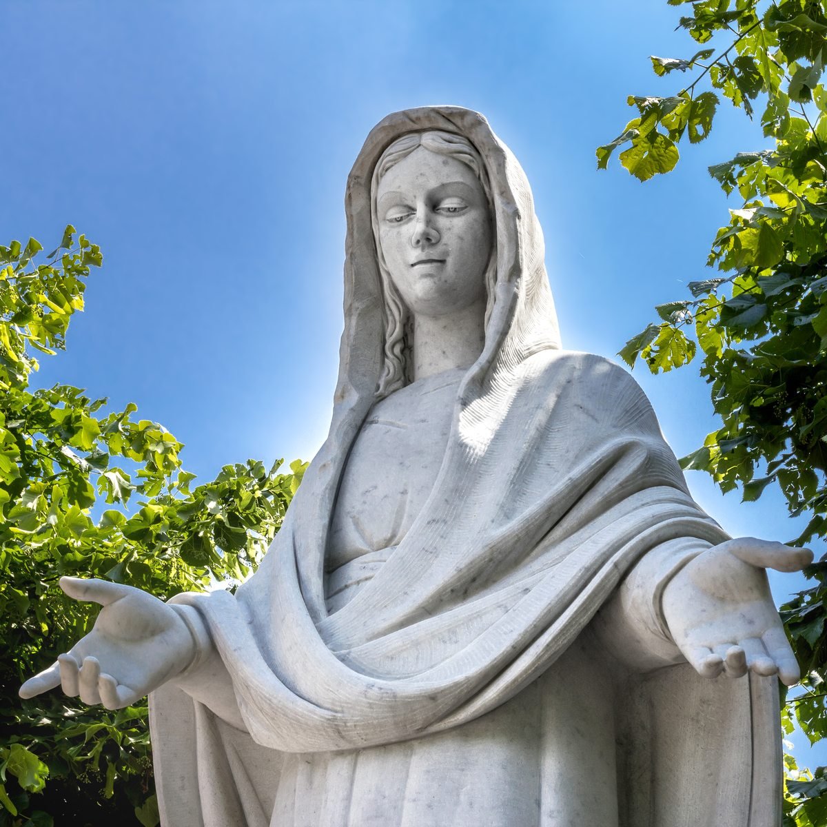 A marble statue of the Blessed Virgin with open hands, against the sky